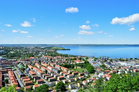 Utsikt över en stad vid en stor sjö med många hus, grönska och blå himmel med moln.