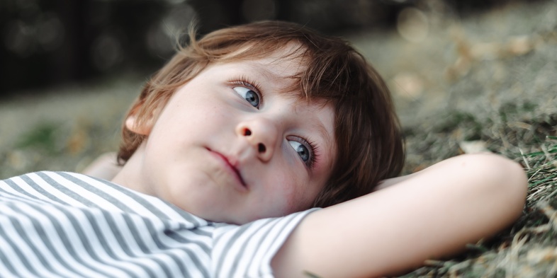 Ett barn med ljusbrunt hår ligger på marken utomhus och tittar drömmande uppåt.