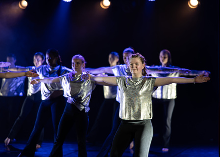 En grupp dansare i silverfärgade tröjor uppträder på scen med utsträckta armar under blått scenljus.
