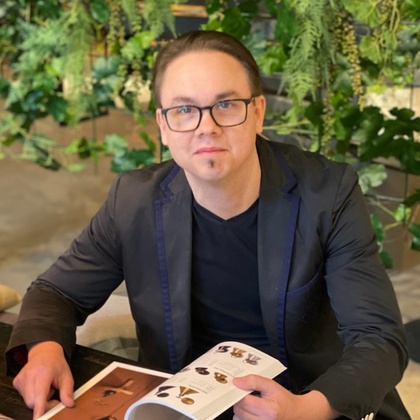 Person i svart kavaj sitter vid ett bord och bläddrar i en tidning, omgiven av gröna växter.