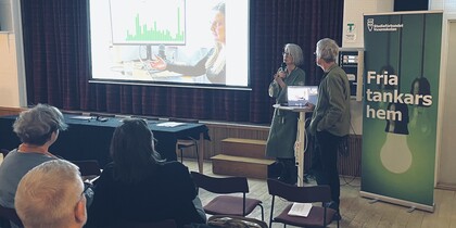 Två personer håller en presentation inför en liten publik med en projektor och en rollup med texten 'Fria tankars hem'.
