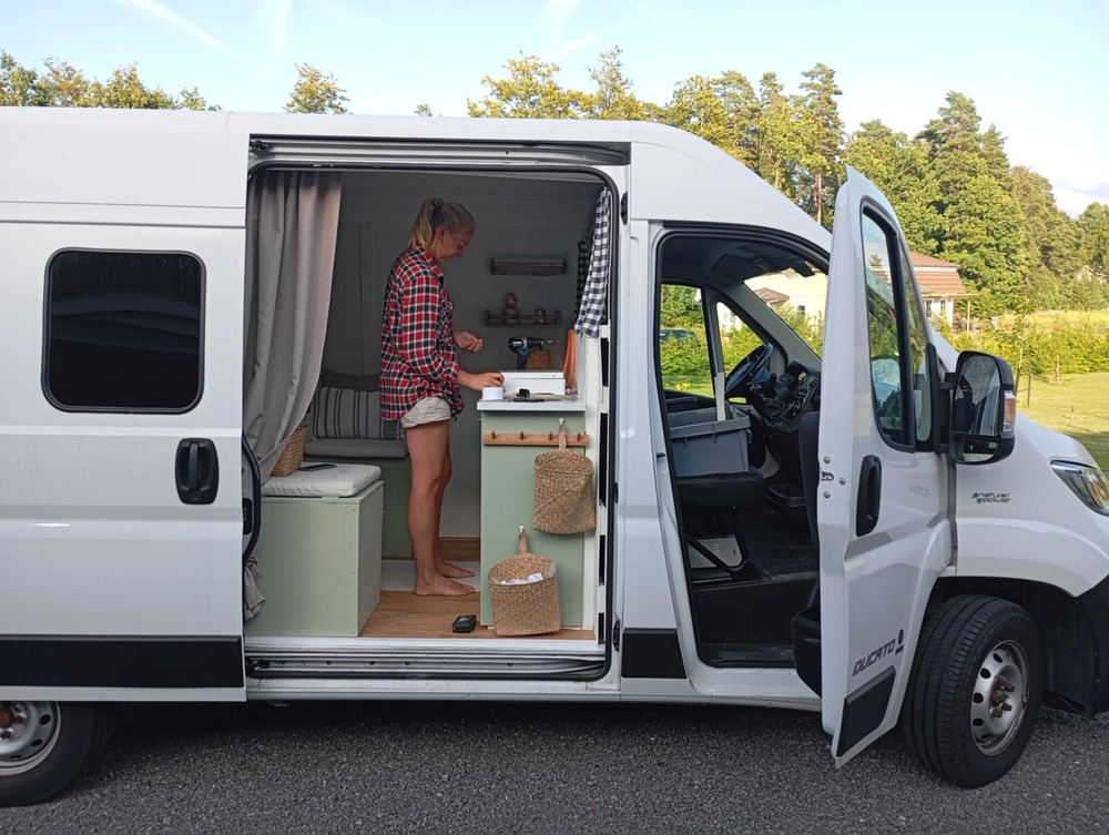 En person står i ett ombyggt skåpbilskök med öppen dörr, parkerad utomhus vid grönska.