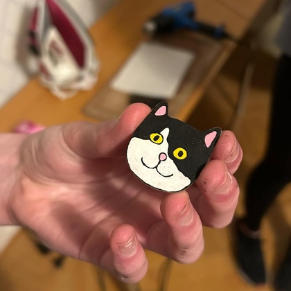 En hand håller en liten figur av en svartvit katt med gula ögon, i ett rum med trägolv och strykjärn i bakgrunden.