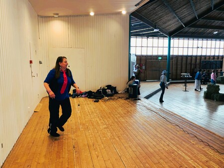 Kvinna i blå skjorta och röd slips står på ett trägolv i en stor ljus lokal med andra personer i bakgrunden.