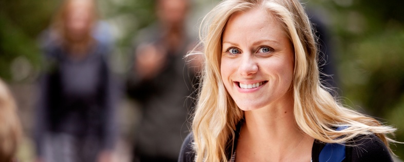 Leende kvinna med blont hår och röd topp står utomhus med suddiga personer i bakgrunden.