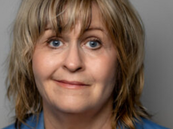 Person med axellångt blont hår, blå kavaj och vit topp mot grå bakgrund.