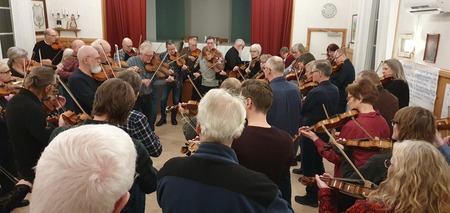 En grupp människor står i en cirkel och spelar fiol tillsammans i ett ljust rum.