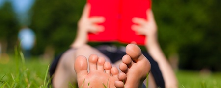 En person ligger på gräset och läser en röd bok, bara fötterna syns tydligt i förgrunden.