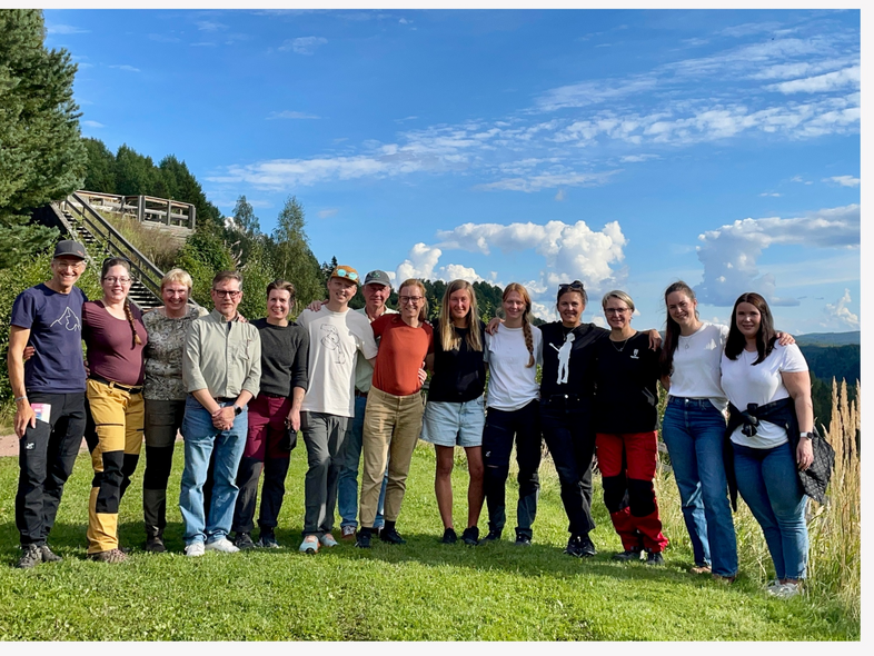 En grupp människor står tillsammans utomhus på en gräsmatta med blå himmel och träd i bakgrunden.