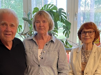 Fyra äldre personer står tillsammans inomhus framför stora fönster och en grön växt.