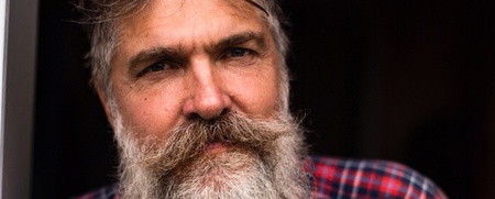 Man med stort grått skägg och glasögon på huvudet, bär rödblå rutig skjorta, står mot mörk bakgrund.