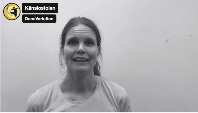 Person med uppsatt hår och ljus tröja sitter mot en vit vägg. Två svarta textskyltar syns i övre vänstra hörnet.