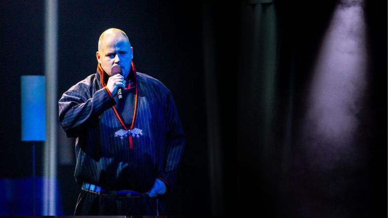 Person står på scen och talar i mikrofon under stark belysning.