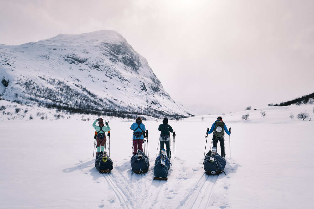 Fyra personer med pulkor står på skidor i ett snötäckt fjällandskap med berg i bakgrunden.