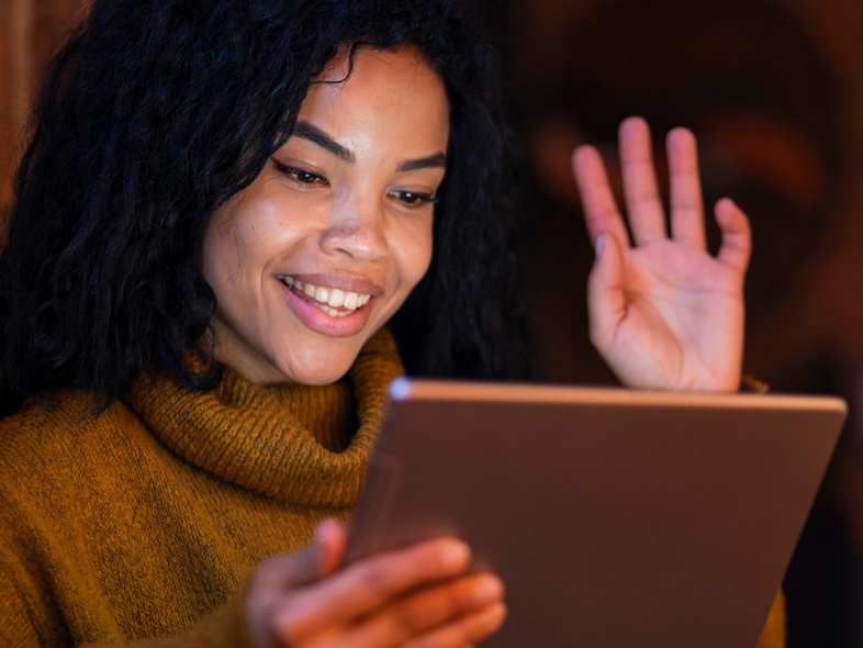 Kvinna med lockigt hår vinkar och ler mot en surfplatta i ett varmt upplyst rum.