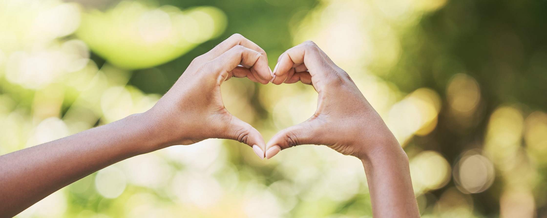 Två händer formar ett hjärta mot en suddig, grön bakgrund utomhus.