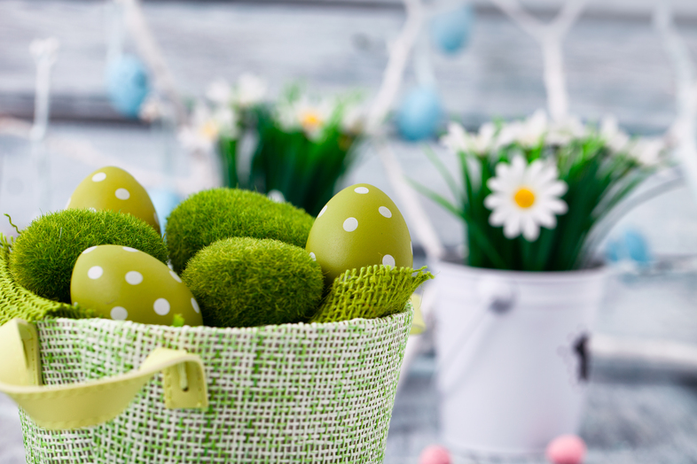Gröna påskägg med vita prickar och konstgräs i en grön korg, med blommor i bakgrunden.