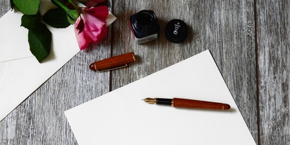 Ett tomt papper, en reservoarpenna, bläckflaska och en ros på ett trägolv, redo för att skriva ett brev.