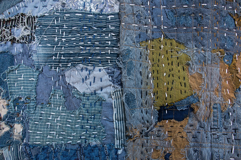 Närbild av lappad textil med synliga stygn i blå, gröna och bruna nyanser.