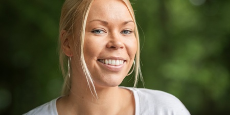 Ung kvinna med blont hår och vit t-shirt ler utomhus i en grönskande miljö.