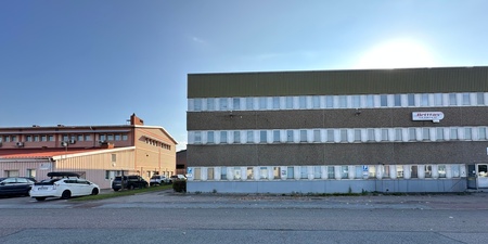 Två industribyggnader med parkerade bilar framför, fotograferade en solig dag.