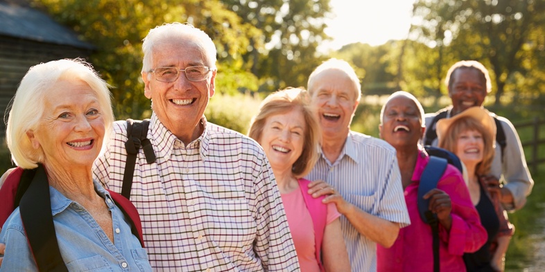 En grupp glada äldre personer står utomhus i rad och ler mot kameran en solig dag.