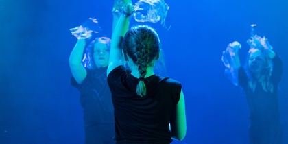 Tre personer i svarta kläder dansar på en scen med blått ljus, en sitter på knä i förgrunden.