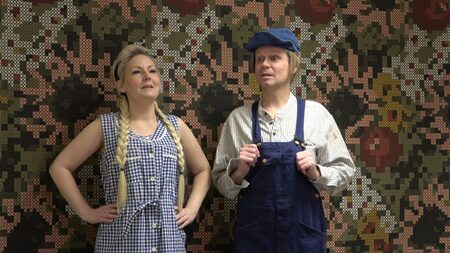 Två personer i rutiga kläder står framför en mönstrad vägg, en kvinna med flätor och en man i blå hängselbyxor och keps.