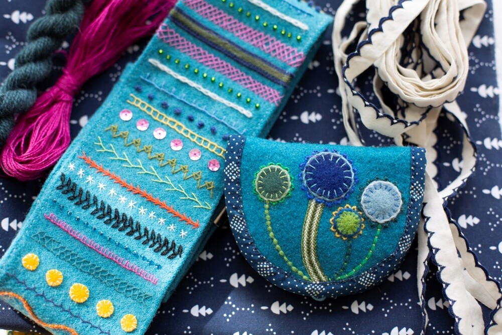Broderade textilier i blå och turkosa nyanser, inklusive en liten väska och band, på ett mönstrat tyg.
