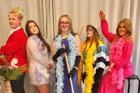 Fem personer i färgglada utklädnader poserar framför ett draperi med rekvisita som fjäderboor, diskokulor och en kvast.