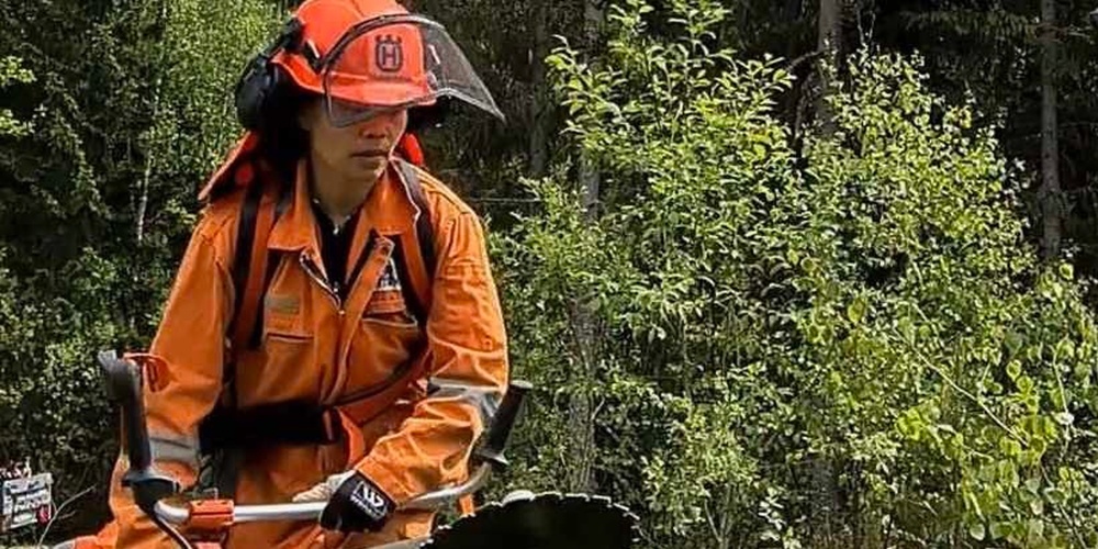 Person i orange skyddskläder arbetar med röjsåg i en skog under blå himmel.