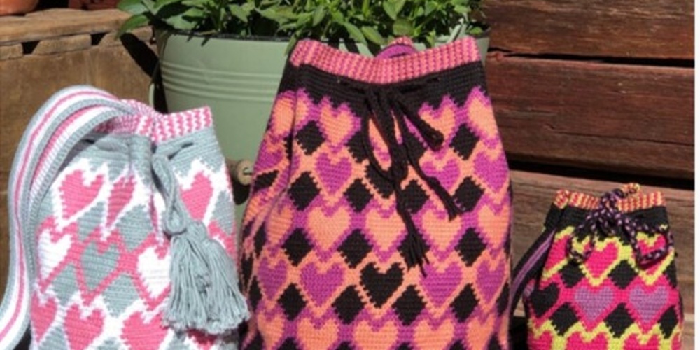 Tre färgglada virkade väskor med hjärtmönster står på en träbänk framför en blomkruka.