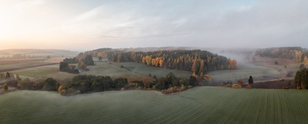 Panoramabild över ett dimmigt landskap med åkrar, skog och morgonljus.
