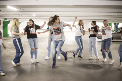 En grupp unga personer dansar tillsammans i ett parkeringsgarage, alla bär jeans och t-shirts med texten 'DANS STUDIO'.