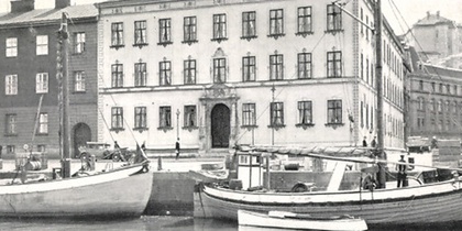 Svartvit bild av båtar vid kajen framför en stor, gammal byggnad i stadsmiljö.