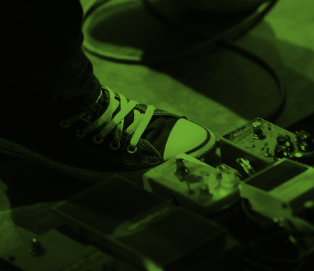 En fot i en svart sneaker trycker på gitarrpedaler på ett golv med grönt ljus.