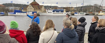 En grupp människor lyssnar på en guide vid en hamn med båtar och byggnader i bakgrunden.