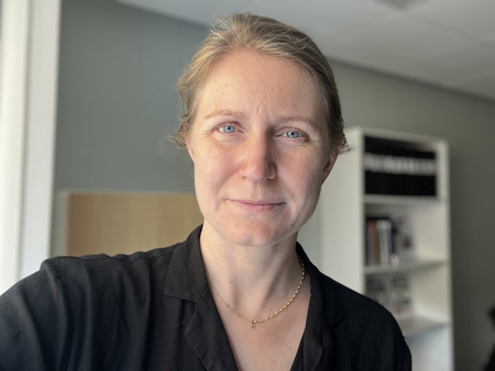 Kvinna med blont hår och svart skjorta står inomhus framför en bokhylla.