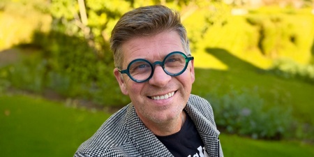 Malte Hallquist i rutigt kavaj och svart t-shirt står utomhus i en grönskande park.