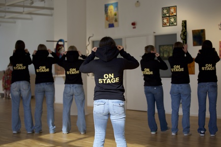 Sju personer står med ryggen mot kameran, alla bär svarta huvtröjor med texten 'ON STAGE' och blå jeans.