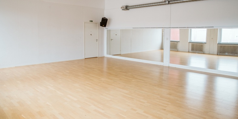 Ljust dans- eller träningsrum med trägolv, stora speglar och vita väggar.