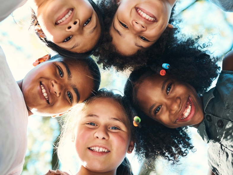 Fem barn står i en cirkel och tittar ner mot kameran, alla ler och ser glada ut utomhus.