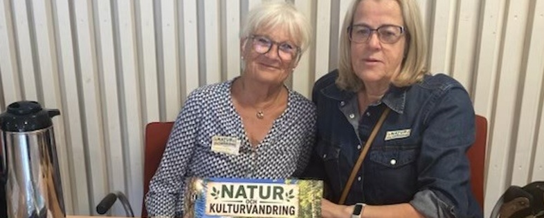 Två kvinnor sitter vid ett bord med material om natur- och kulturvandring, dekorerat med naturföremål och informationsskyltar.