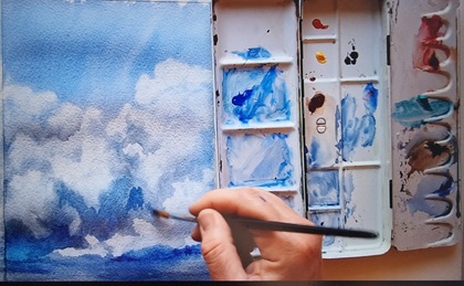 En hand målar moln på ett papper med akvarellfärger och pensel, färgpalett syns bredvid.