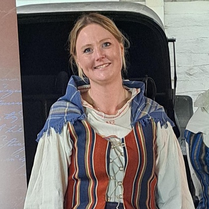 Kvinna i traditionell folkdräkt med randig väst och kjol står inomhus och ler mot kameran.