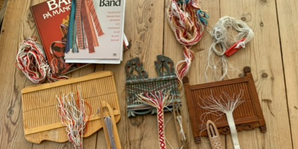 Bandvävning med olika redskap och garn på ett träbord, samt böcker om bandvävning.