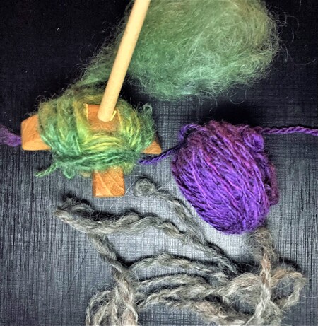 Färgad ull, en träslända och garnnystan i grönt, lila och grått på svart bakgrund.