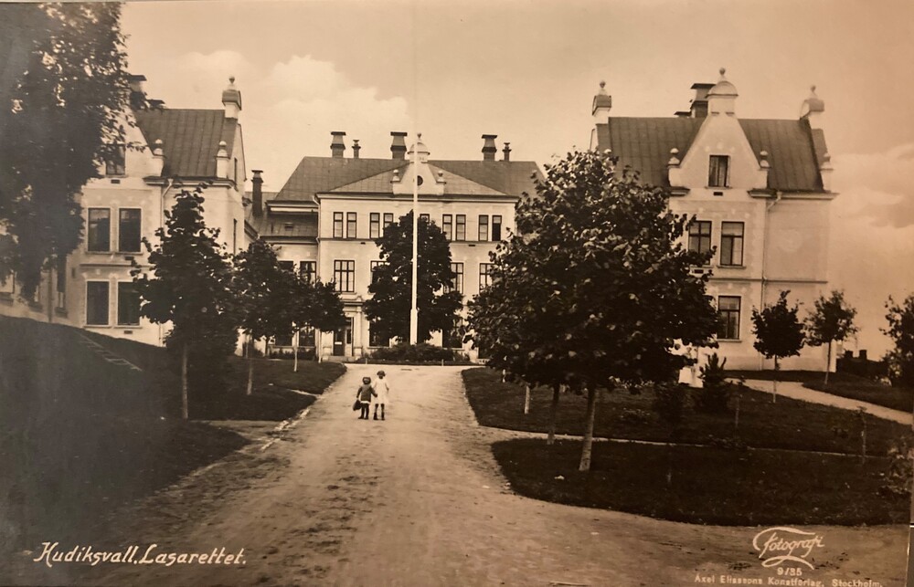 Gammalt svartvitt foto av Hudiksvalls lasarett med två barn som går på en grusväg mot byggnaden.