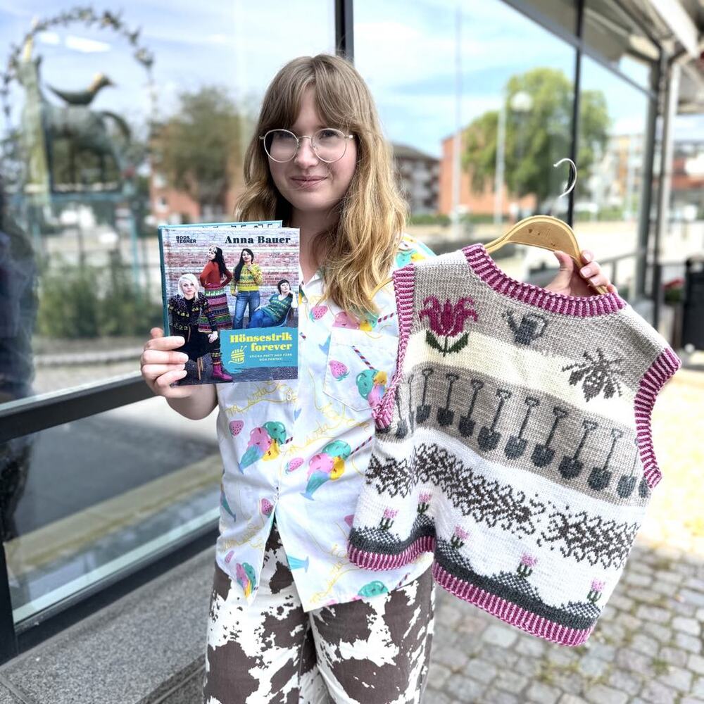 Person håller upp en stickad väst med trädgårdsmotiv och en bok utanför en byggnad.