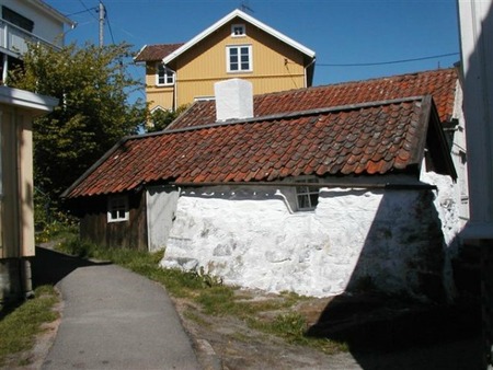 Vitkalkat stenhus med rött tegeltak vid en smal asfalterad gång, omgiven av trähus i solljus.